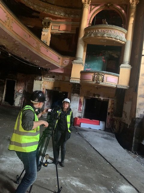 Two people inside old Empire Theatre in Burnley wearing High visibility Vests and Hard hats while one operates a video camera filming the interview with Sophie Gibson